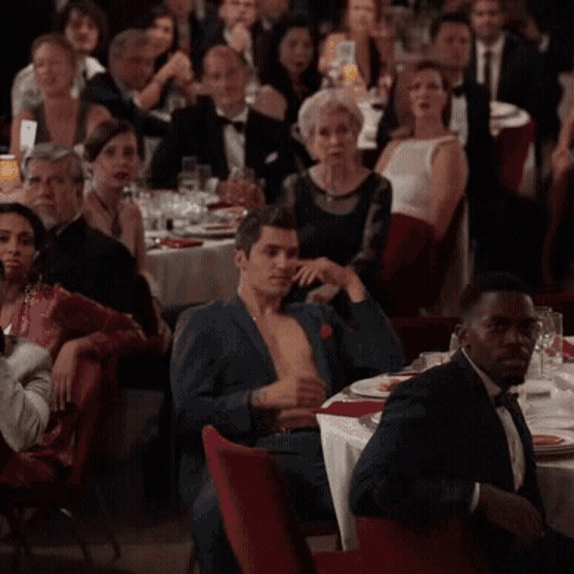 a man without a shirt is sitting in front of a crowd of people at a dinner party .