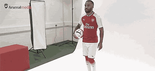 a man in a fly emirates shirt is holding a soccer ball in a room .