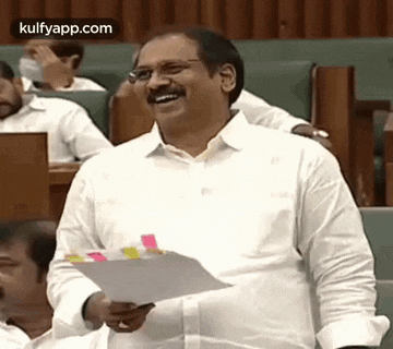 a man in a white shirt is laughing while holding a piece of paper in a classroom .