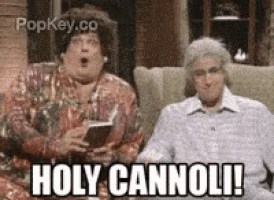 two women are sitting on a couch and one of them is holding a book and the other is saying holy cannoli !