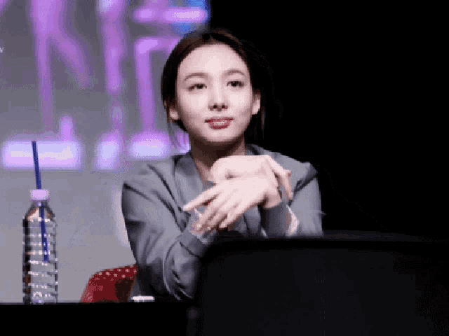 a woman is sitting at a table with her hands folded and a bottle of water next to her .