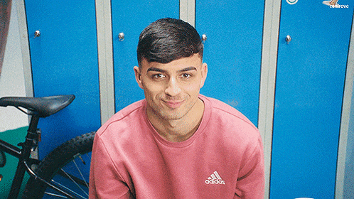 a man wearing a pink adidas sweatshirt is sitting in front of blue lockers