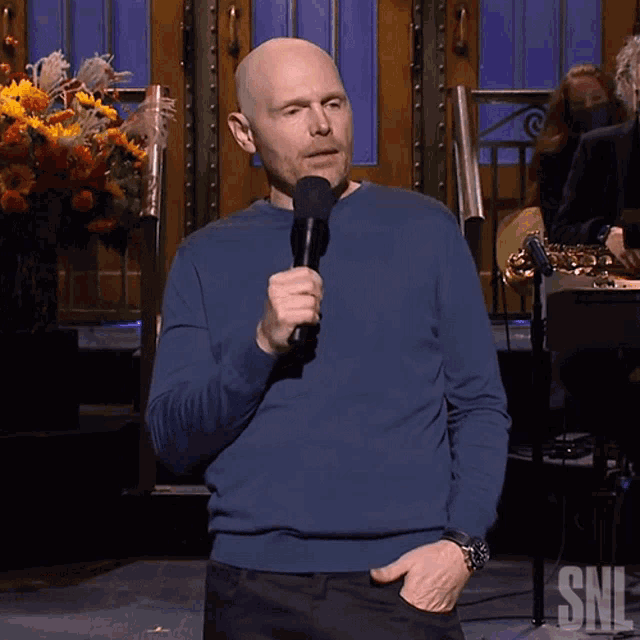 a man in a blue sweater is holding a microphone with a snl logo in the corner