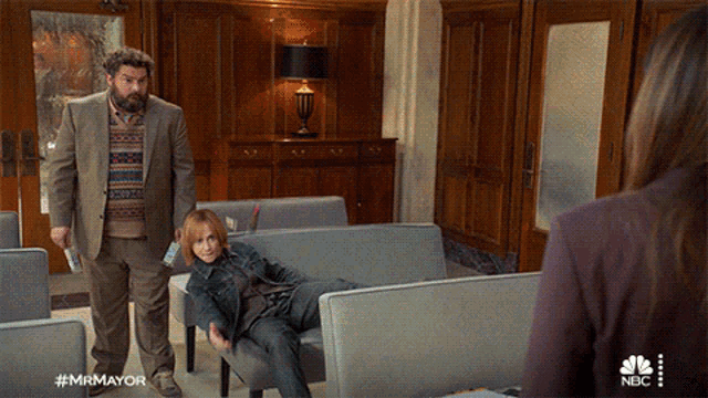 a man in a suit stands next to a woman laying on a couch in a waiting room