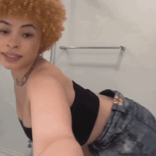 a woman with curly hair is taking a selfie in a bathroom while wearing a black bra and denim shorts .