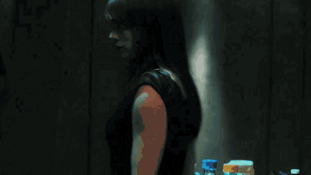 a woman in a black dress stands in a dark room with bottles in the background