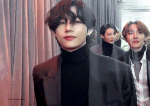 a man in a black suit and turtleneck is standing in front of a group of men in suits .