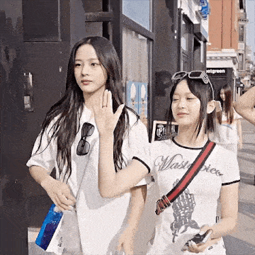 two young women are walking down a street and one of them is wearing a t-shirt that says wastepiece .