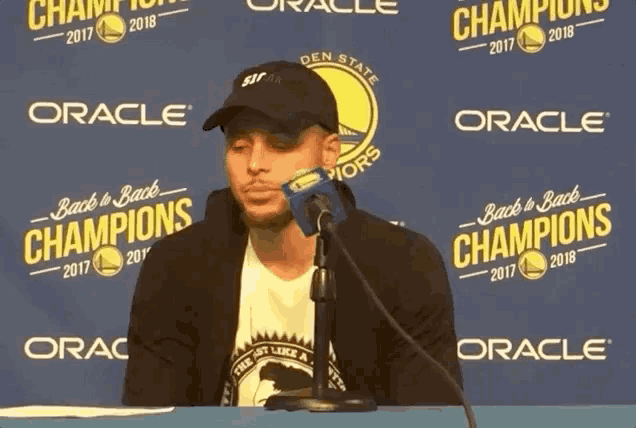 a man sitting at a table with a microphone in front of a back to back champions banner