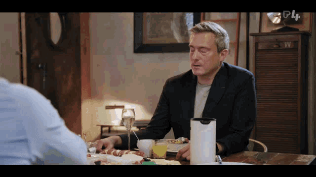 a man in a suit sits at a table with a cup of orange juice in front of him