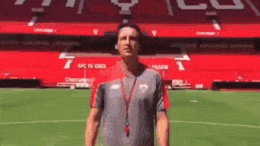a man in a grey shirt is standing on a soccer field in front of a stadium .
