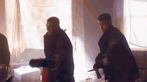 two firefighters are standing next to each other in a room holding a fire extinguisher and a knife .