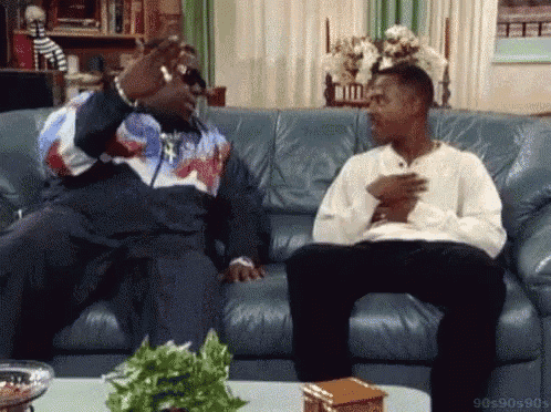 two men are sitting on a couch in a living room talking .