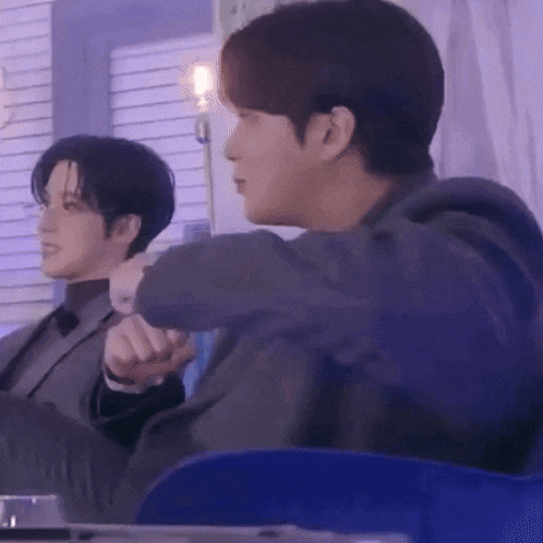 a man in a suit is sitting next to another man in a blue chair and holding his hand out .