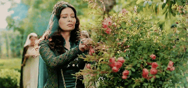 a woman in a green dress is standing next to a bush of roses .