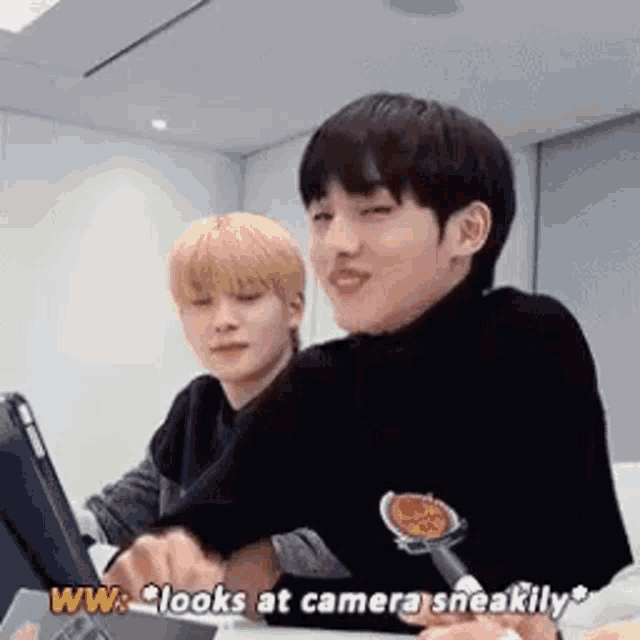 two young men are sitting at a table looking at a tablet and one of them is saying `` looks at camera sneakily '' .