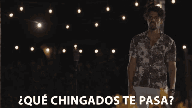 a man stands in front of a string of lights with the words " qué chingados te pasa " above him