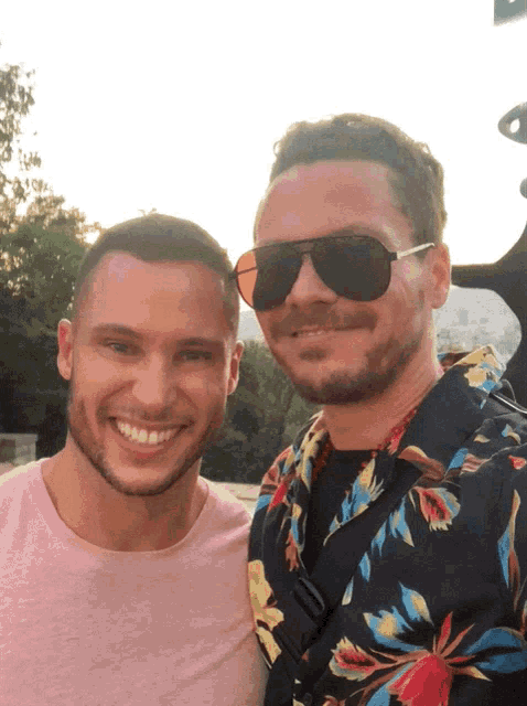 a man wearing sunglasses stands next to another man in a floral shirt
