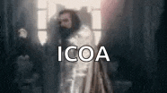 a man with a beard is standing in front of a window in a dark room with the word icoa on the bottom .
