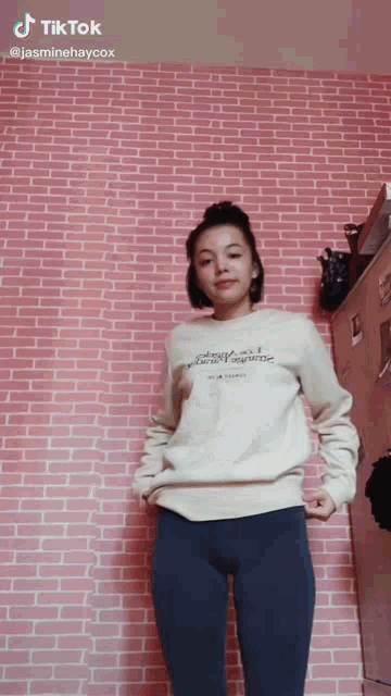 a woman is standing in front of a pink brick wall wearing a white shirt and blue leggings .