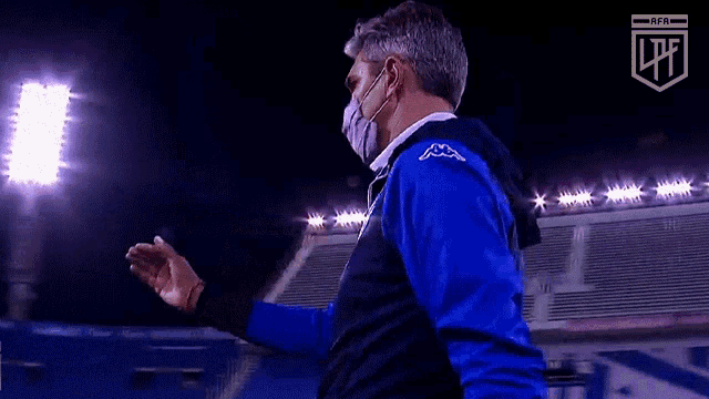 a man wearing a mask and a blue kappa jacket stands on a soccer field .