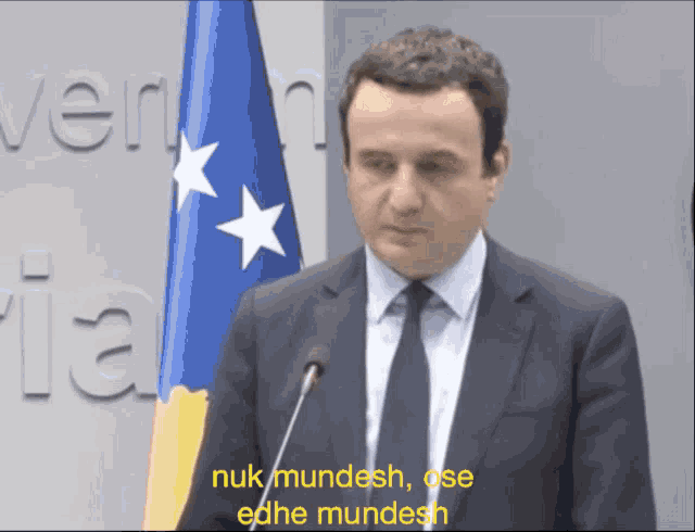 a man in a suit and tie stands in front of a microphone with the words nuk mundesh on the screen behind him