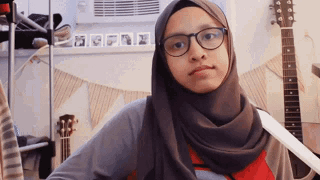 a woman wearing glasses and a hijab is standing in front of a guitar