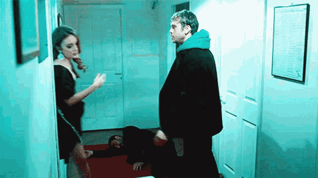 a man in a blue hoodie stands next to a woman in a black dress in a hallway