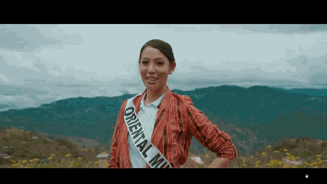 a woman wearing a oriental miss sash stands in front of mountains