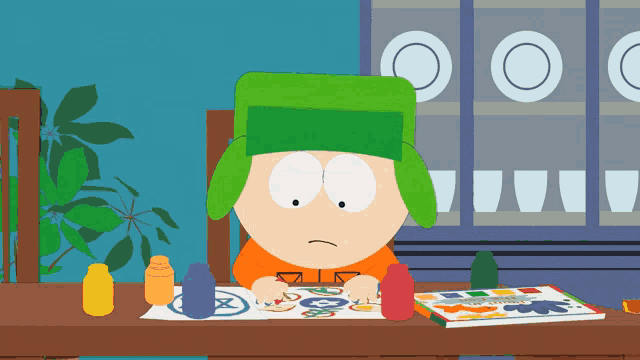 a boy in a green hat sits at a table with crayons