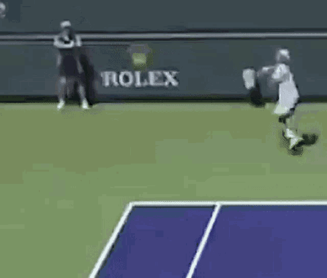 a couple of people standing on top of a tennis court next to a wall that says rolex .