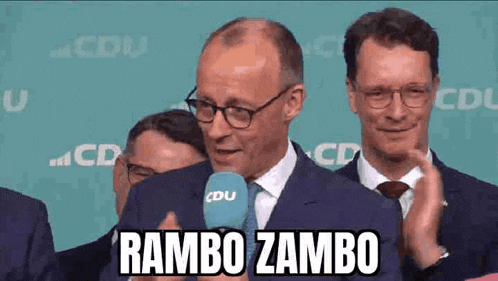 a man in a suit and tie is talking into a microphone and says rambo zambo .