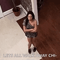 a woman in a dress is standing in a hallway with her arms crossed and says `` lets all walk away chi '' .