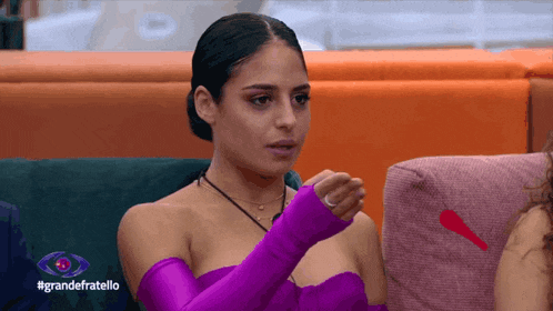 a woman in a purple dress is sitting on a couch with the hashtag grandefratello on the bottom