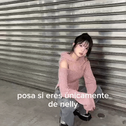 a woman in a pink sweater and jeans is sitting on the ground in front of a metal shutter .