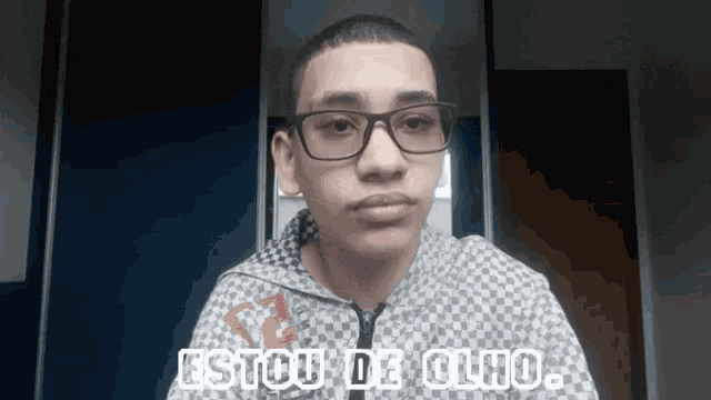 a young man wearing glasses and a checkered jacket says " estou de olho "