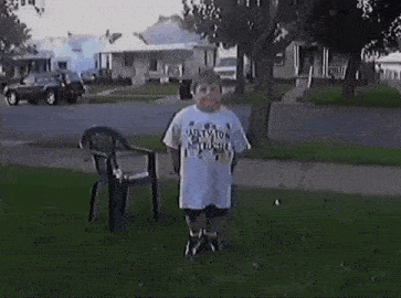 a boy in a white shirt is jumping in the grass next to a chair .