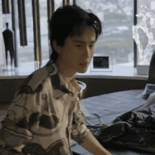 a man is sitting on a bed in front of a window looking at something .