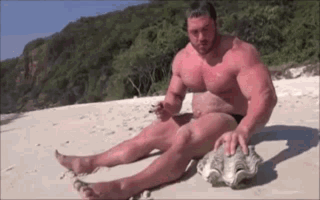 a shirtless man is sitting on a beach holding a large shell .