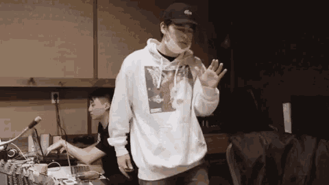 a man wearing a white hoodie and a black hat is standing in a recording studio .