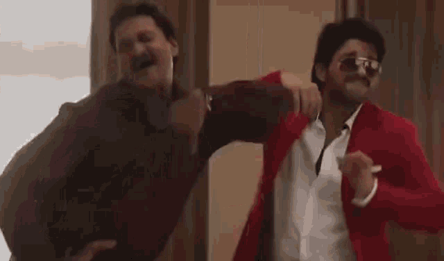 two men are dancing together in a room . one of the men is wearing a red jacket .