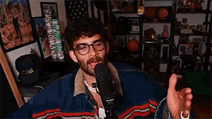a man wearing glasses and a pearl necklace is talking into a microphone in a room .