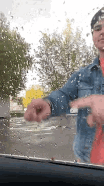 a man in a denim jacket is standing in front of a car windshield with rain drops on it .