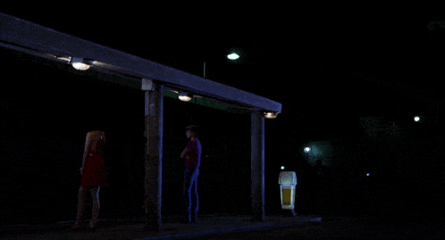 a woman in a red dress stands next to a woman in a blue shirt at a bus stop