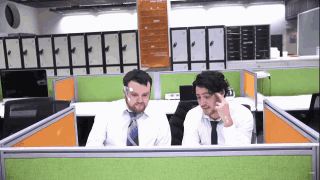 two men sit at a desk in an office cubicle looking at a computer monitor