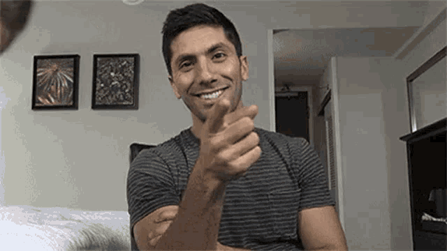 a man is smiling and pointing at the camera while sitting on a bed in a bedroom .