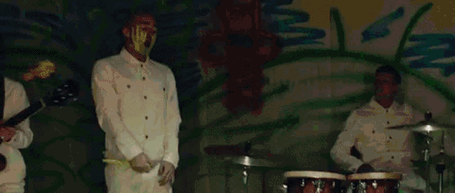 a man with a yellow mask on his face is playing drums in front of a wall with graffiti on it .