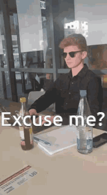 a man wearing sunglasses sits at a table with a bottle of water and a notebook .
