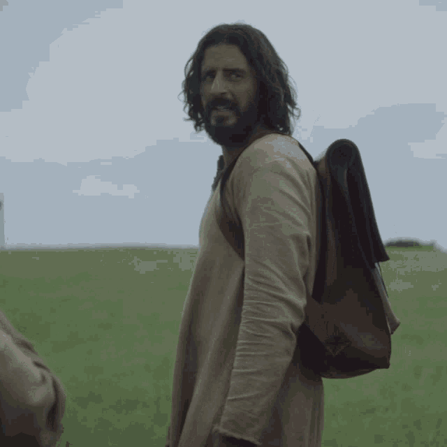 a man with a beard and a backpack stands in a grassy field