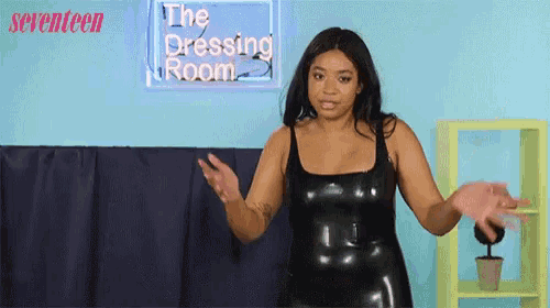 a woman in a black latex dress stands in front of a neon sign that says the dressing room